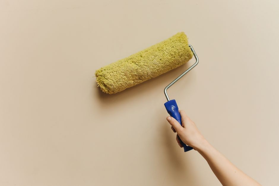 A hand using a yellow paint roller on a beige wall.